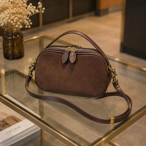 May include: A dark brown suede handbag with a rounded rectangular shape, a top handle, and a detachable shoulder strap. The bag features a gold-toned zipper closure with two matching suede pull tabs and gold metal hardware attachments on the sides.