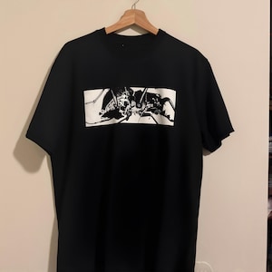 May include: A black short-sleeved t-shirt featuring a rectangular white graphic print on the chest. The graphic displays a detailed black and white illustration of a mechanical insect-like creature with sharp appendages and thin wire-like lines.