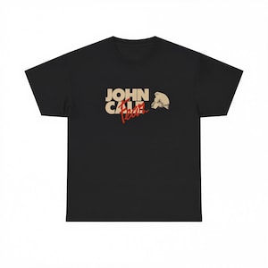 Könnte beinhalten: Ein schwarzes Kurzarm-T-Shirt mit einem Grafikdruck auf der Brust. Die Grafik zeigt den Text JOHN CALE in großen beigen Buchstaben, darunter das Wort Fear in roter Schreibschrift sowie eine kleine beige Illustration einer menschlichen Figur.