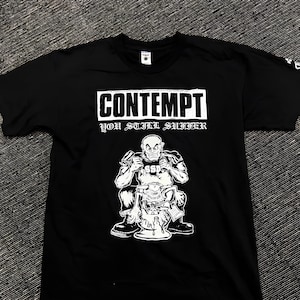 Può includere: Una t-shirt nera a maniche corte con una stampa grafica bianca. Il design include il testo CONTEMPT e YOU STILL SUFFER sopra un'illustrazione di una figura calva seduta su un water, con le lettere SSD stampate sulla maglietta della figura.