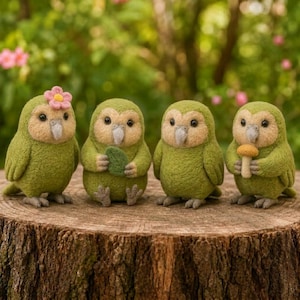 May include: Four needle-felted green owl figurines with beige faces and grey beaks. The first owl wears a pink flower, the second holds a green leaf, and the fourth holds a small brown mushroom. They are arranged in a row on a textured tree stump.