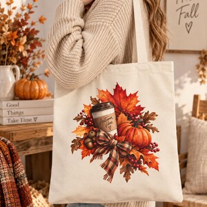 Könnte beinhalten: Eine cremefarbene Stofftasche aus Segeltuch mit einem aufgedruckten Motiv eines Kaffeebechers mit der Aufschrift Autumn Vibes, umgeben von orangefarbenen und roten Ahornblättern, einem Kürbis, Eicheln, roten Beeren und einer karierten Schleife.
