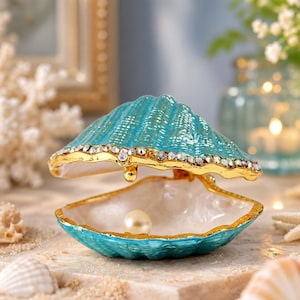 May include: A turquoise seashell-shaped trinket box with a gold-tone rim encrusted with clear crystals. The hinged lid is open, revealing a smooth white interior and a single, centered, round white pearl resting inside the base.