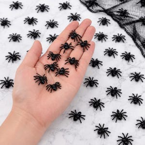 Peut inclure: Une collection de petites figurines d'araignées en plastique noir dispersées sur une surface à motif de marbre blanc, avec plusieurs araignées reposant dans une paume ouverte et un morceau de filet décoratif noir visible dans le coin supérieur droit.