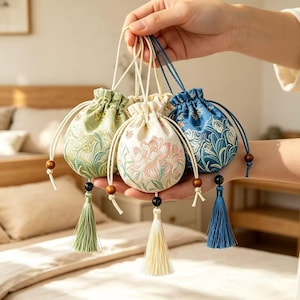 May include: Three small drawstring pouches in light green, cream, and blue, each featuring intricate floral embroidery. The pouches have matching fabric drawstrings with wooden and black beads, finished with long, dangling silk tassels at the base.