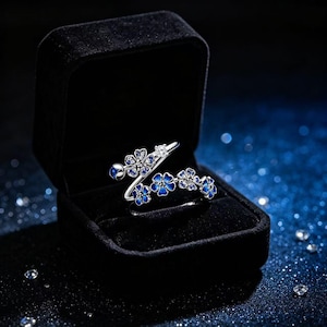 May include: A silver-toned open ring featuring a series of small, five-petaled flowers with vibrant blue enamel centers and clear, sparkling stones. The ring is displayed inside a black velvet jewelry box with a hinged lid and a soft interior.