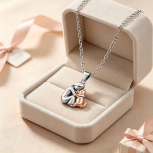 Könnte beinhalten: Eine silberfarbene Metall-Halskette mit einem Anhänger, der eine stilisierte Figur zeigt, die ein kleines Tier umarmt. Die Figur ist poliert silberfarben, während das Tier in Roségold gehalten ist. Der Anhänger hängt an einer feinen Kette.