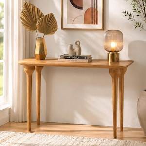 Può includere: Un tavolo console in legno marrone chiaro con quattro gambe affusolate, dotato di un vaso dorato con due foglie di palma dorate, una piccola scultura astratta beige, due libri impilati con la scritta Art Deco e una lampada in vetro.