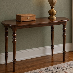 May include: A dark brown wooden console table with an oval top and four turned legs. A rectangular wooden box with a gold-colored metal latch sits on the left side of the table, and a brass-toned table lamp stands on the right side.