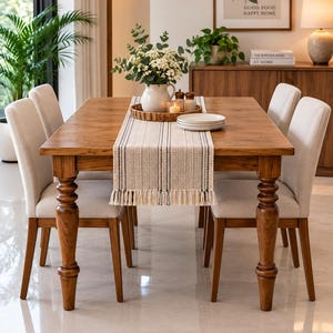 May include: A rectangular wooden dining table with four upholstered chairs featuring light beige fabric. A woven table runner with black and white stripes and fringe ends sits on the table, topped with a white pitcher of flowers and plates.