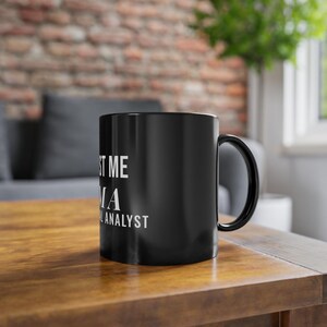 Pode incluir: Uma caneca de cerâmica preta com alça, apresentando um texto impresso em branco que diz TRUST ME I AM A FINANCIAL ANALYST. A caneca tem um acabamento liso e brilhante e uma forma cilíndrica, projetada para conter bebidas quentes ou frias.