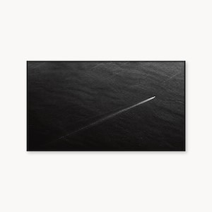 Puede incluir: Una fotografía aérea en blanco y negro que muestra un pequeño y delgado barco blanco moviéndose a través de una vasta y oscura extensión de agua. La embarcación deja una estela blanca, larga, recta y diagonal que corta la superficie texturizada del mar.