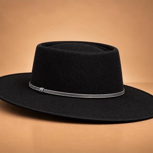 May include: A black wide-brimmed hat with a flat crown and a stiff, structured silhouette. The hat features a thin, decorative silver-toned cord band wrapped around the base of the crown, secured with a small, intricate knot detail at the front.