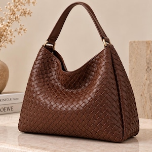 Può includere: Una borsa hobo marrone scuro caratterizzata da un intricato motivo in pelle intrecciata su tutto l'esterno. La borsa presenta un unico manico arcuato fissato con anelli in metallo dorato, una silhouette morbida e un'ampia apertura superiore.