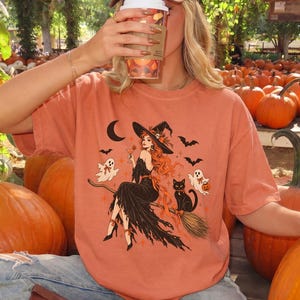 May include: A burnt orange t-shirt featuring a graphic print of a witch with long red hair sitting on a broomstick, accompanied by a black cat, two ghosts, bats, and a crescent moon. The shirt is worn with blue jeans and brown boots.