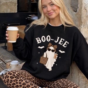 May include: A black crewneck sweatshirt featuring a graphic of a ghost wearing sunglasses and a leopard print bow, holding a leopard print tote bag and a coffee cup. The text BOO-JEE is printed above the ghost, accompanied by small bat and star icons.