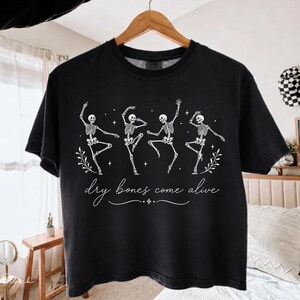 May include: A black short-sleeve t-shirt featuring a white graphic of four dancing skeletons surrounded by small stars and leaf sprigs. Below the skeletons, the text reads "dry bones come alive" in a white cursive script font.