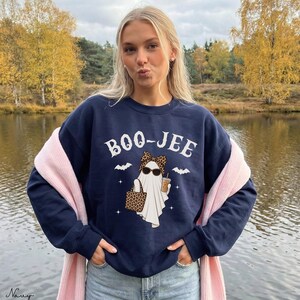 May include: A navy blue crewneck sweatshirt featuring a white graphic of a ghost wearing a leopard print bow and sunglasses, holding a leopard print tote bag and a coffee cup. The text BOO-JEE is printed above the ghost with small bats and stars.
