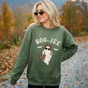 May include: An olive green crewneck sweatshirt featuring a graphic of a white ghost wearing a leopard print bow and sunglasses, holding a leopard print tote bag and a coffee cup. The text BOO-JEE is printed above the ghost with small bats.