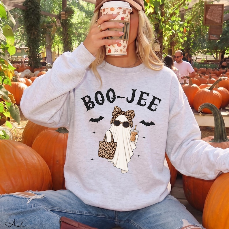 May include: A light gray crewneck sweatshirt featuring a graphic of a ghost wearing a leopard print bow and sunglasses, holding a leopard print tote bag and a coffee cup. The text BOO-JEE is printed above the ghost with small bat icons.