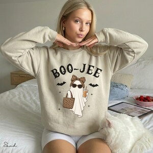 May include: A sand-colored crewneck sweatshirt featuring a graphic of a ghost wearing a leopard print bow and sunglasses, holding a leopard print tote bag and a coffee cup. The text BOO-JEE is printed above the ghost, flanked by two black bats.