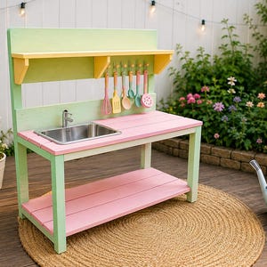 May include: A wooden play kitchen featuring a light green frame, a pink countertop, and a pink lower shelf. It includes a stainless steel sink with a faucet, a yellow shelf above, and six colorful plastic kitchen utensils hanging on hooks.