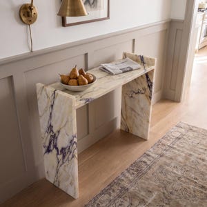 May include: A rectangular console table crafted from white marble with prominent purple and gold veining. The table features a flat top surface supported by two vertical slab legs. A white bowl filled with brown pears sits on the table.