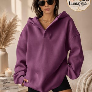 Könnte beinhalten: Ein dunkelvioletter Oversize-Kapuzenpullover mit weitem Kragen, langen Ärmeln mit gerippten Bündchen und einer Vordertasche. Das Kleidungsstück wird mit einer schwarzen Shorts getragen. Der Kapuzenpullover hat eine lockere Passform mit überschnittenen Schultern.