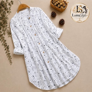 May include: A white long-sleeve tunic shirt featuring a small black floral print, a button-down front with light brown buttons, a mandarin collar, and pleated detailing along the chest. The shirt is displayed on a wooden hanger.