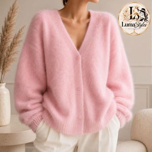 Puede incluir: Un cárdigan de color rosa claro con cuello en V, suave y esponjoso, con mangas largas y tres botones a juego en la parte delantera. El tejido de punto tiene un aspecto texturizado y cepillado. La prenda se lleva sobre pantalones blancos.