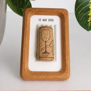 May include: A rectangular wooden frame containing a cork with a wine glass graphic and the text 12 AUG 2026 and 95087 printed on it. The cork is mounted on a white background with a light green striped pattern behind the cork inside the frame.
