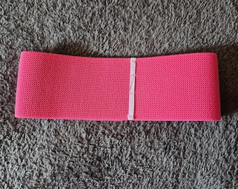 May include: A bright pink fabric resistance band featuring a textured, woven surface and a single vertical white stitched seam running through the center of the band. The band is rectangular in shape with straight edges and a consistent width throughout.
