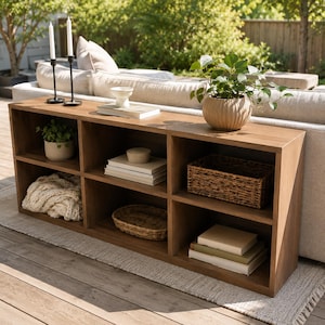 Puede incluir: Una mesa consola rectangular de madera con seis compartimentos de almacenamiento abiertos. La superficie superior sostiene dos portavelas negros con velas blancas, un cuenco blanco pequeño sobre un libro y una planta en una maceta de cerámica beige.