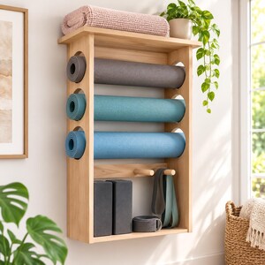 May include: A light-coloured wooden wall-mounted storage rack featuring a top shelf with a folded pink towel, three circular cutouts holding grey, teal, and blue yoga mats, two wooden pegs, two grey foam blocks, and two hanging yoga straps.