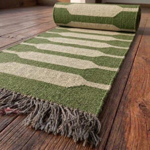 May include: A rectangular woven runner rug featuring a repeating pattern of olive green and cream-colored geometric shapes. The rug is partially rolled at one end and finished with a fringe of loose, textured fibers along the bottom edge.
