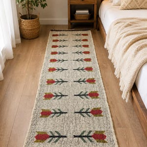 May include: A long, rectangular runner rug featuring a cream-colored base with a repeating pattern of red and green tulip-like floral motifs arranged in two vertical columns. The rug has dark gray fringe detailing at both ends of the textile.