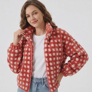 May include: A fuzzy, long-sleeved zip-up jacket featuring a red and white checkered pattern. The jacket has a high collar, a full-length front zipper, and a soft, textured fleece-like material. It is worn over a plain white t-shirt and blue jeans.