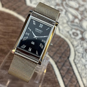May include: A rectangular Seiko Quartz watch with a black dial, silver-tone Roman numeral hour markers, and silver hands. The watch features a polished silver-tone metal case and a fine gold-tone mesh metal strap with a sliding clasp mechanism.