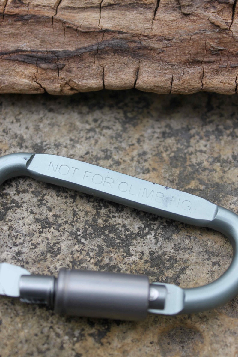 May include: A silver-colored carabiner with the text "NOT FOR CLIMBING" engraved on the side. The carabiner is set against a textured, gray stone surface and a piece of weathered wood.