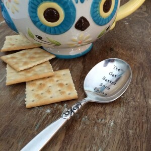 May include: A silver spoon with the words "The Get Better Soon Spoon" engraved on the bowl. The spoon is lying on a wooden surface next to a bowl of soup and crackers.