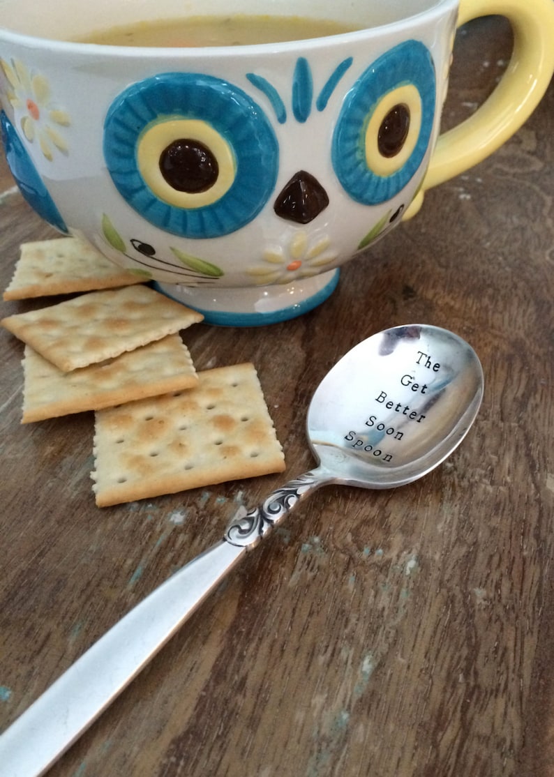 May include: A silver spoon with the words "The Get Better Soon Spoon" engraved on the bowl. The spoon is lying on a wooden surface next to a bowl of soup and crackers.