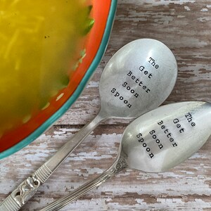 May include: Two silver spoons with the words "The Get Better Soon Spoon" engraved on them. The spoons are laying on a wooden surface.