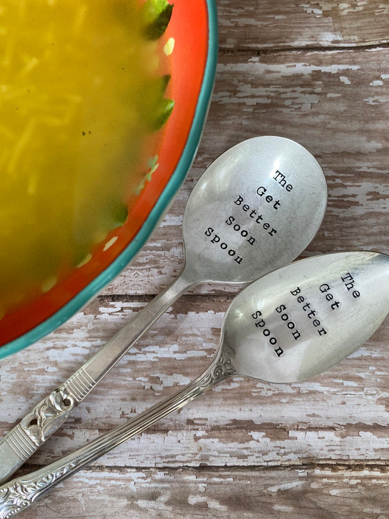 May include: Two silver spoons with the words "The Get Better Soon Spoon" engraved on them. The spoons are laying on a wooden surface.