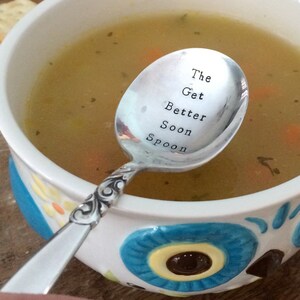 May include: A silver spoon with the words "The Get Better Soon Spoon" engraved on it. The spoon is resting in a bowl of chicken soup.