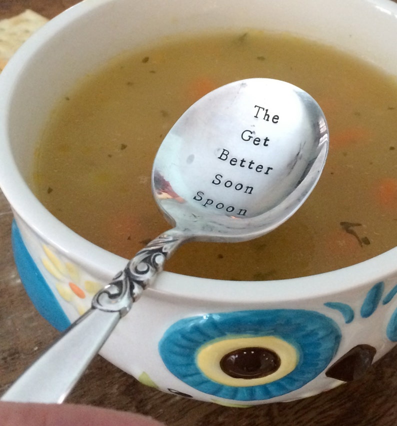 May include: A silver spoon with the words "The Get Better Soon Spoon" engraved on it. The spoon is resting in a bowl of chicken soup.