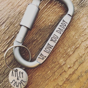 May include: A silver carabiner keychain with a round tag and a key ring. The carabiner is engraved with "WE LOVE YOU DADDY". The round tag is engraved with "ATLEY & SAWYER". The keychain is on a wooden surface.