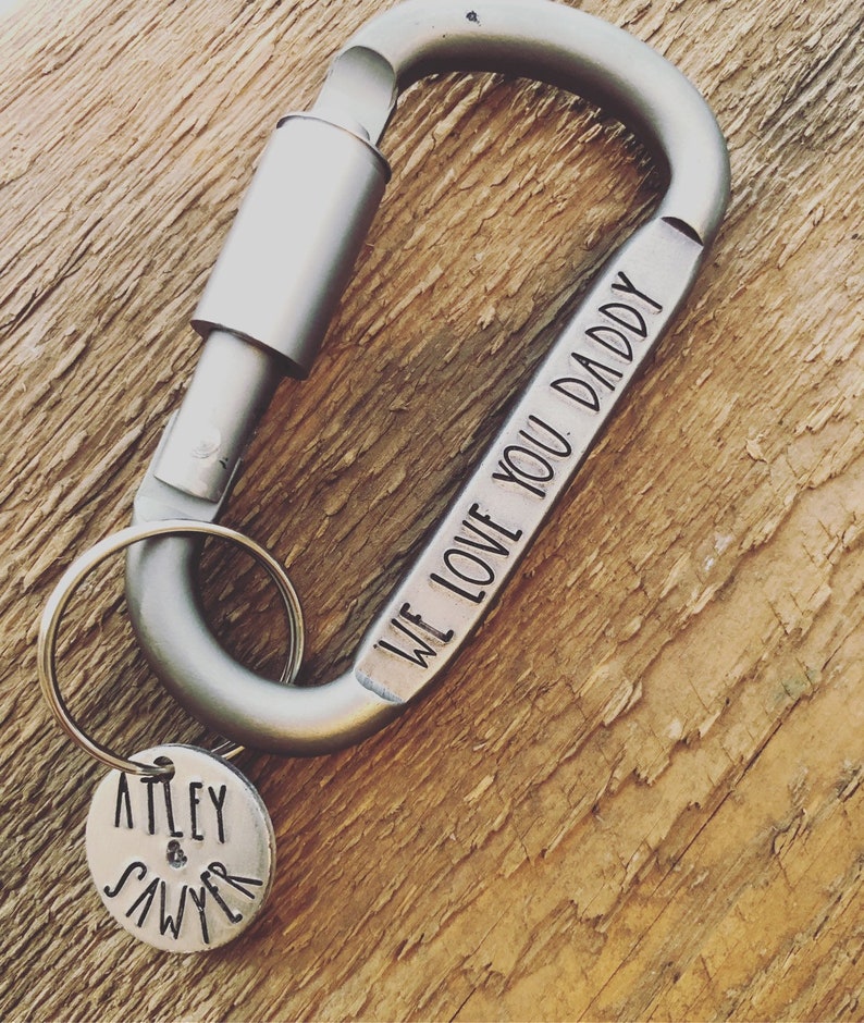 May include: A silver carabiner keychain with a round tag and a key ring. The carabiner is engraved with "WE LOVE YOU DADDY". The round tag is engraved with "ATLEY & SAWYER". The keychain is on a wooden surface.