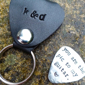 May include: A black leather guitar pick holder with a silver ring and the initials "K & D" engraved on the front. A silver guitar pick with the text "You are the pic to my guitar." is also shown.