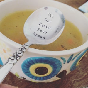 May include: A silver spoon with the words "The Get Better Soon Spoon" engraved on it. The spoon is resting in a white bowl with a blue and yellow owl design. The bowl contains a yellow broth.