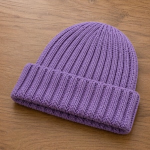 May include: A vibrant lavender beanie crafted from a thick, chunky knit yarn. This hat features a prominent vertical ribbed texture throughout the crown and a wide, folded cuff at the base, providing a structured and consistent appearance.